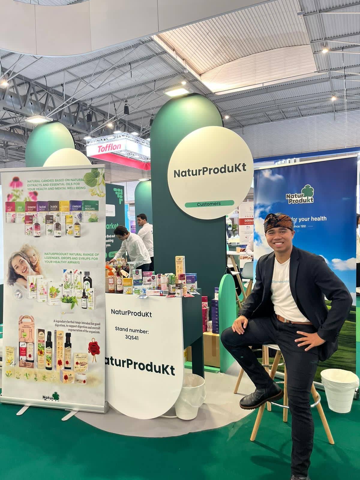 Export manažer sedí u stánku NaturProdukt na veletrhu, zelené a modré bannery, produkty a plakáty v hale.