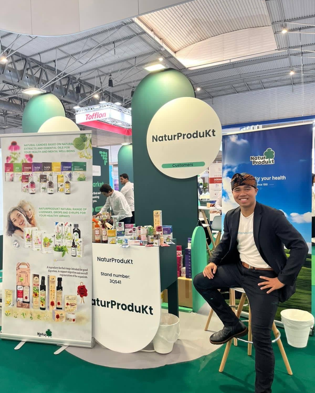 Muž sedí u stánku NaturProdukt na veletrhu, kolem jsou produkty a zelené dekorativní kruhy.