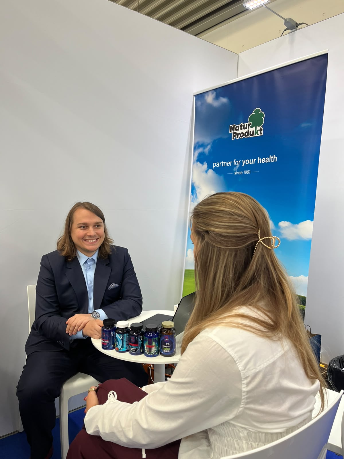 Naturprodukt CEO sedí u stánku, žena mu podává ruku; na stole jsou doplňky.
