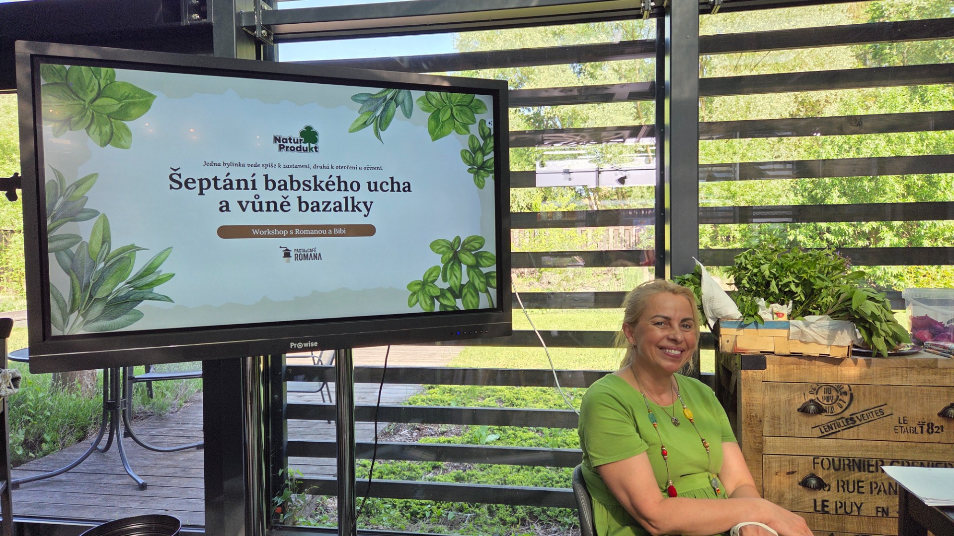 Usměvavá žena v zeleném tričku sedí u monitoru s prezentací o bylinkách a bazalce.