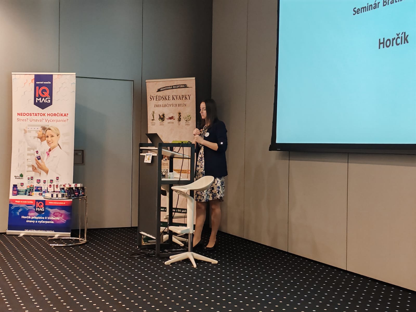 Žena před stojanem s letáky a prezentací, vedle roll-up banneru v konferenční místnosti.