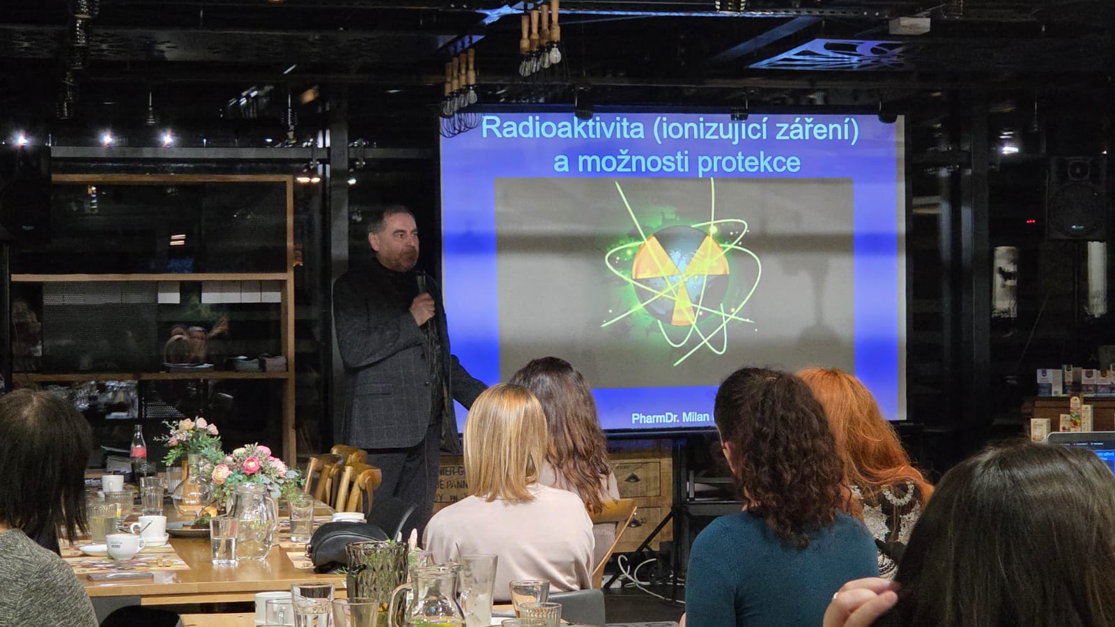 Přednáška v sále, řečník u stolu, promítané téma radioaktivita a ochrana, publikum sedí.