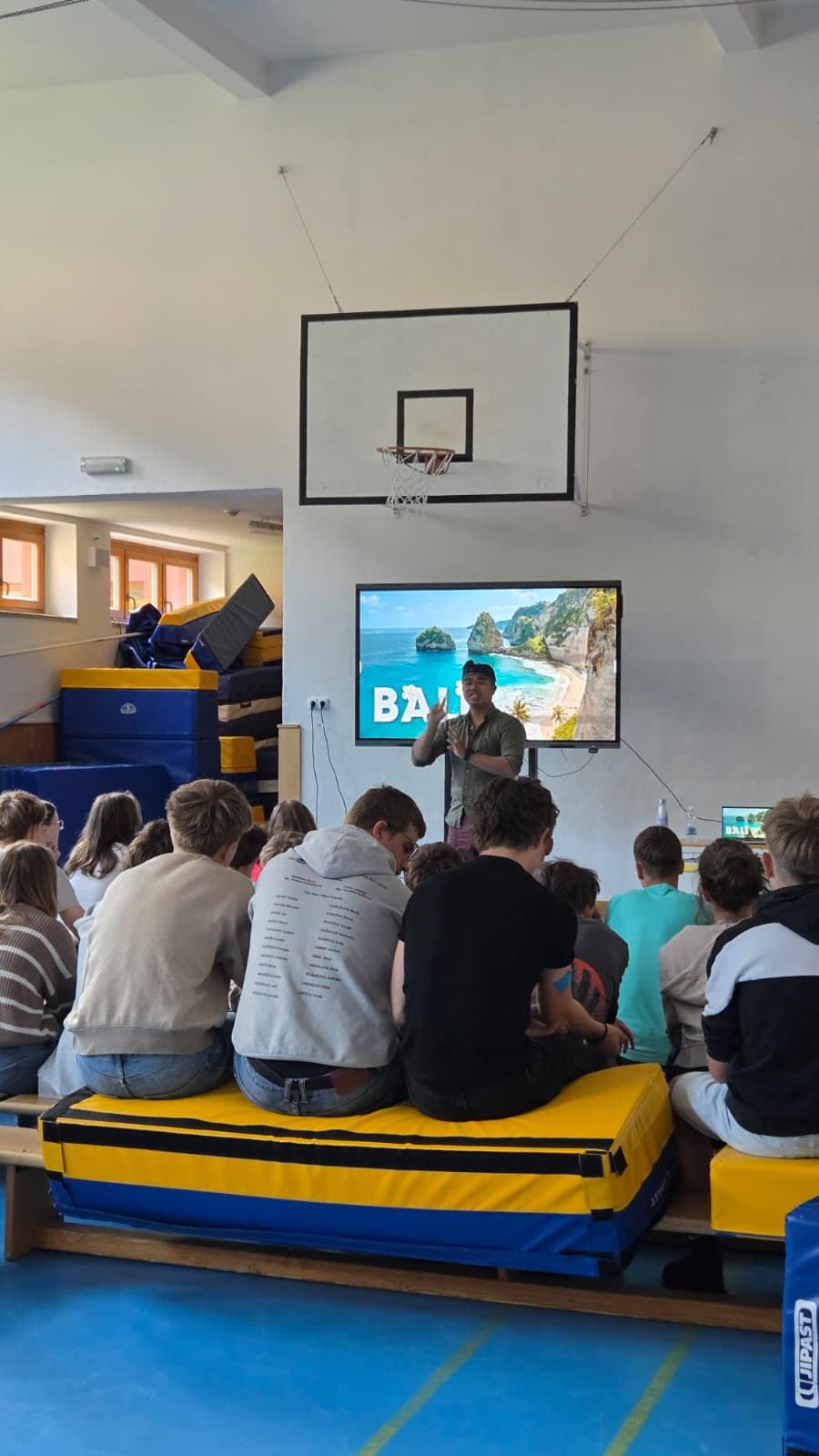 Instruktor přednáší před dětmi v tělocvičně, promítá se obraz „BALI“ na velké obrazovce.