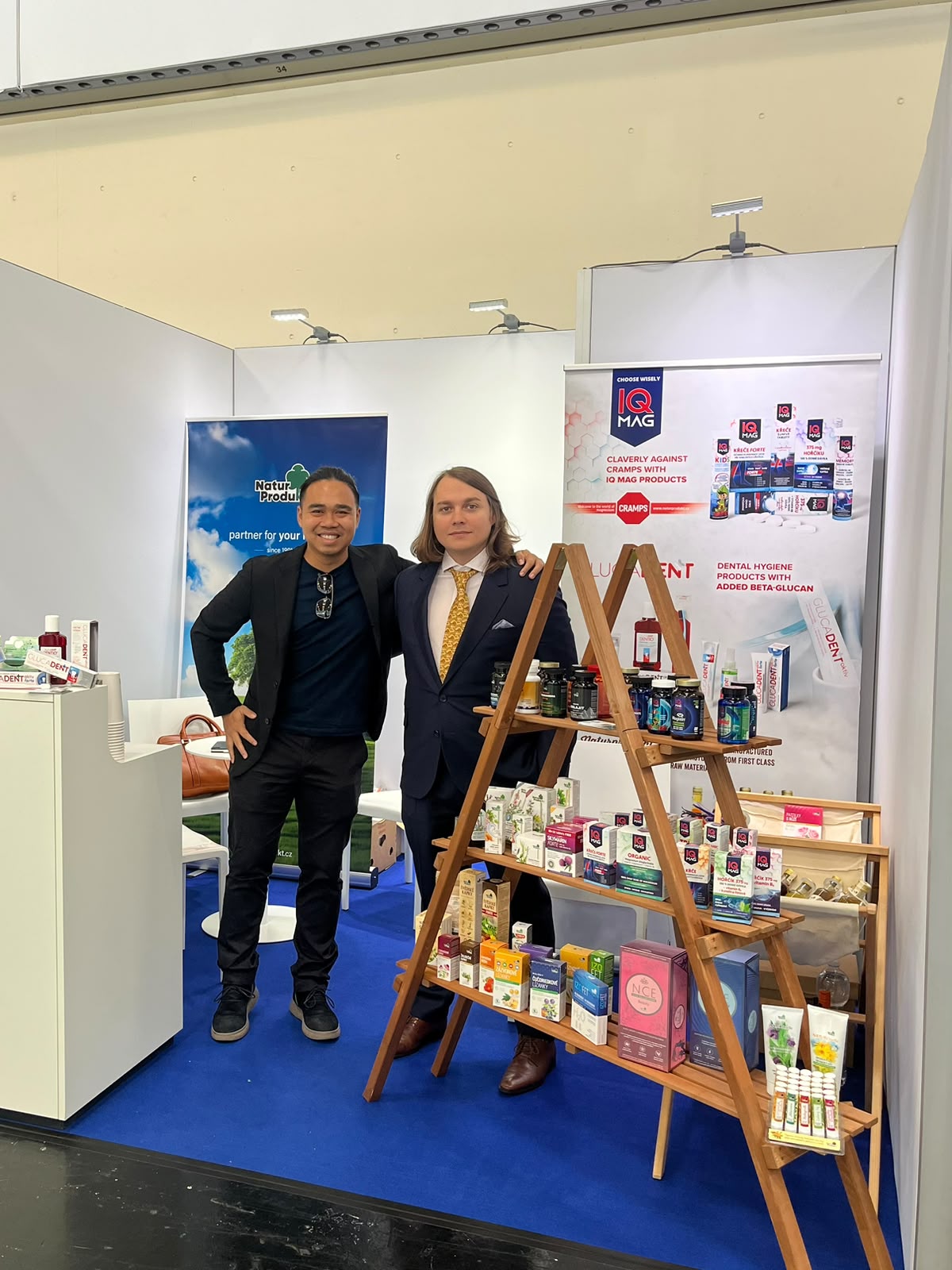 Naturprodukt export manažer a CEO v oblecích stojí na veletrhu u stojanu s produkty IQ MAG a plakáty.